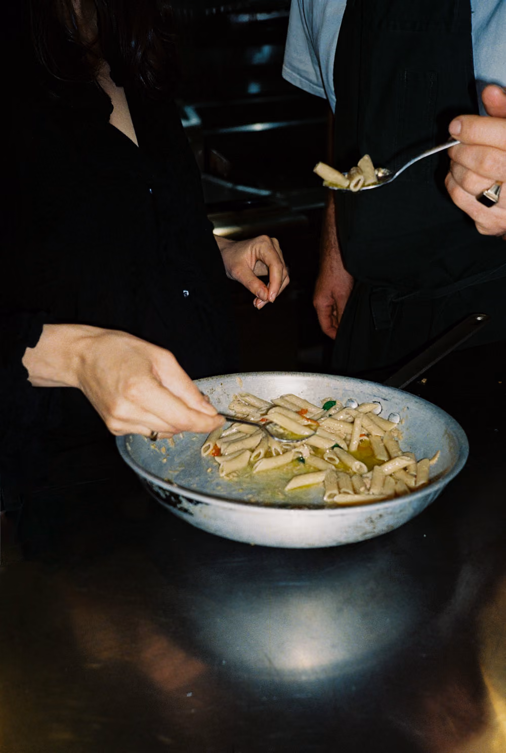 Two people tasting General Wholefoods pasta from a large pan with spoons in a kitchen setting.