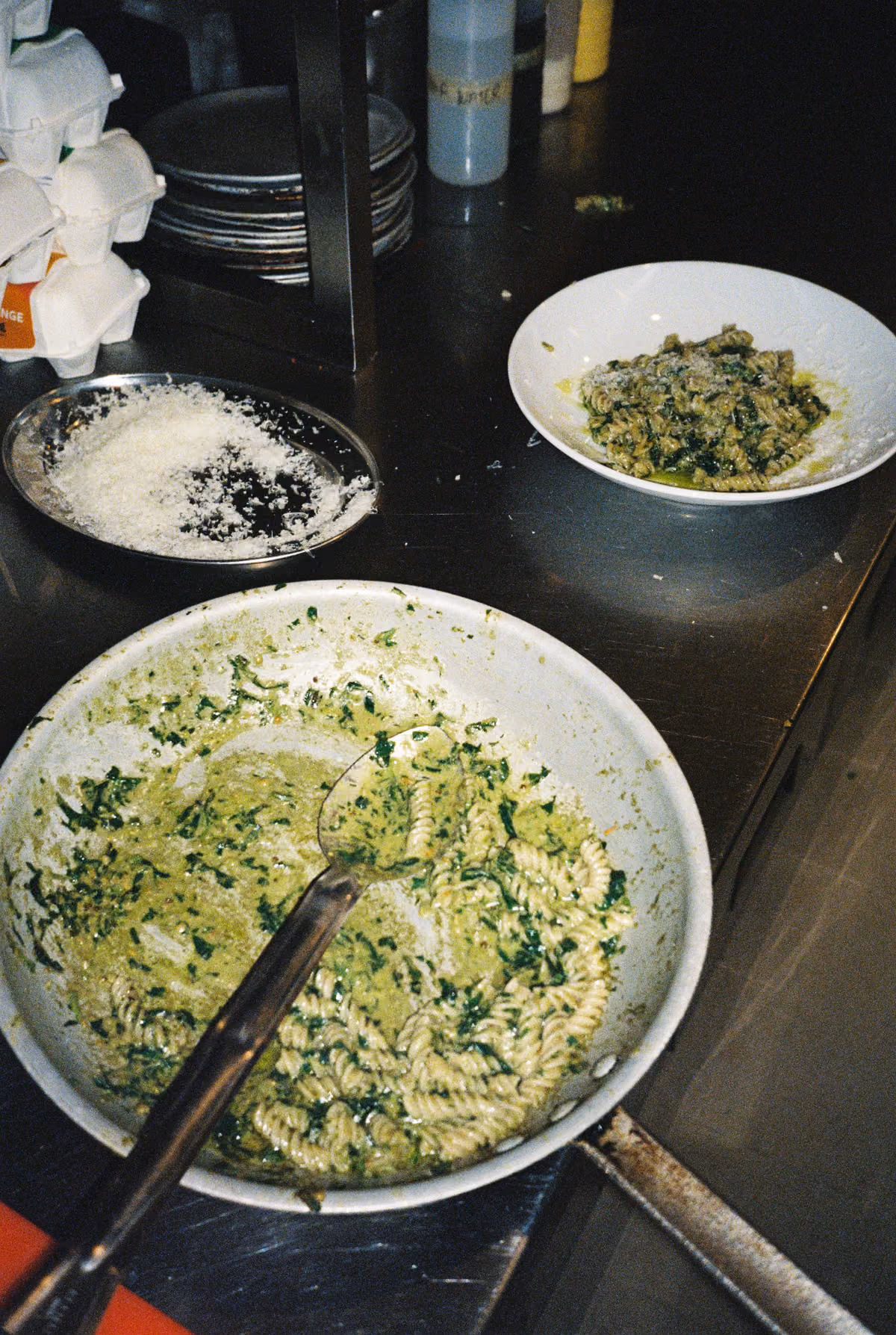 Two bowls of fusilli pasta with green pesto sauce on a metal countertop, one large bowl with a spoon and one smaller bowl in the background, along with a plate of grated cheese and stacks of plates nearby.