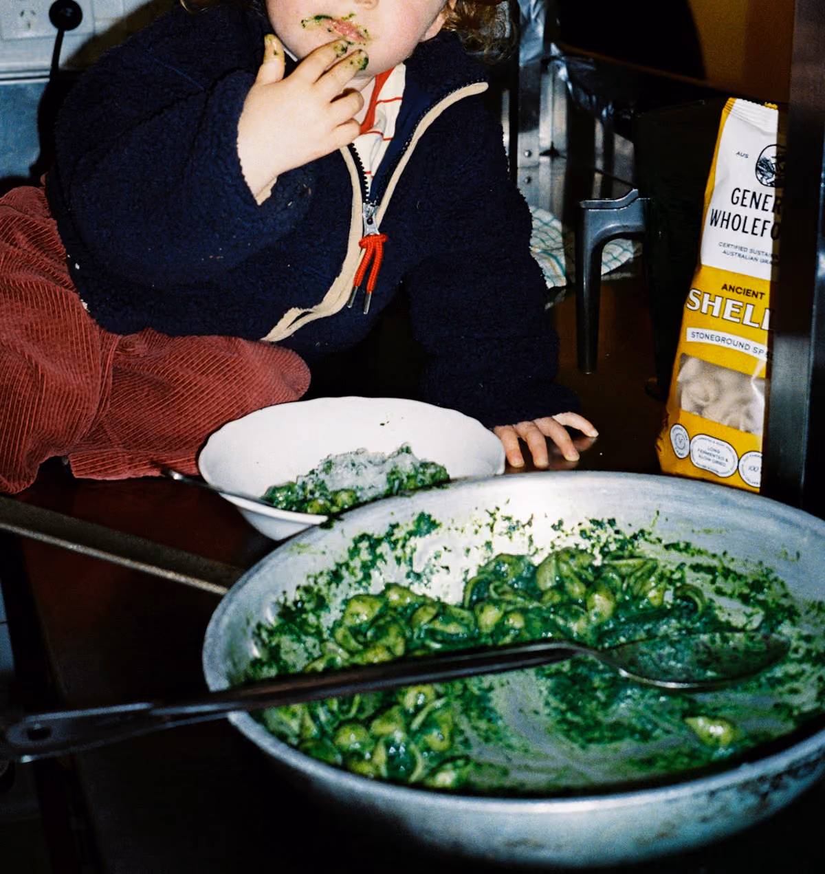 A young child with red pants and a dark jacket eating green pasta with their hands from a white bowl, sitting on a dark surface next to a larger bowl of green pasta and a bag of ancient shell pasta.