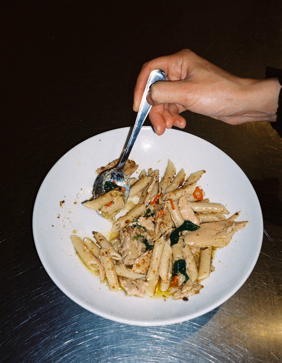 White plate with penne pasta mixed with pieces of tuna, green leafy herbs, and small red pepper bits, held by a hand gripping a spoon scooping the pasta.