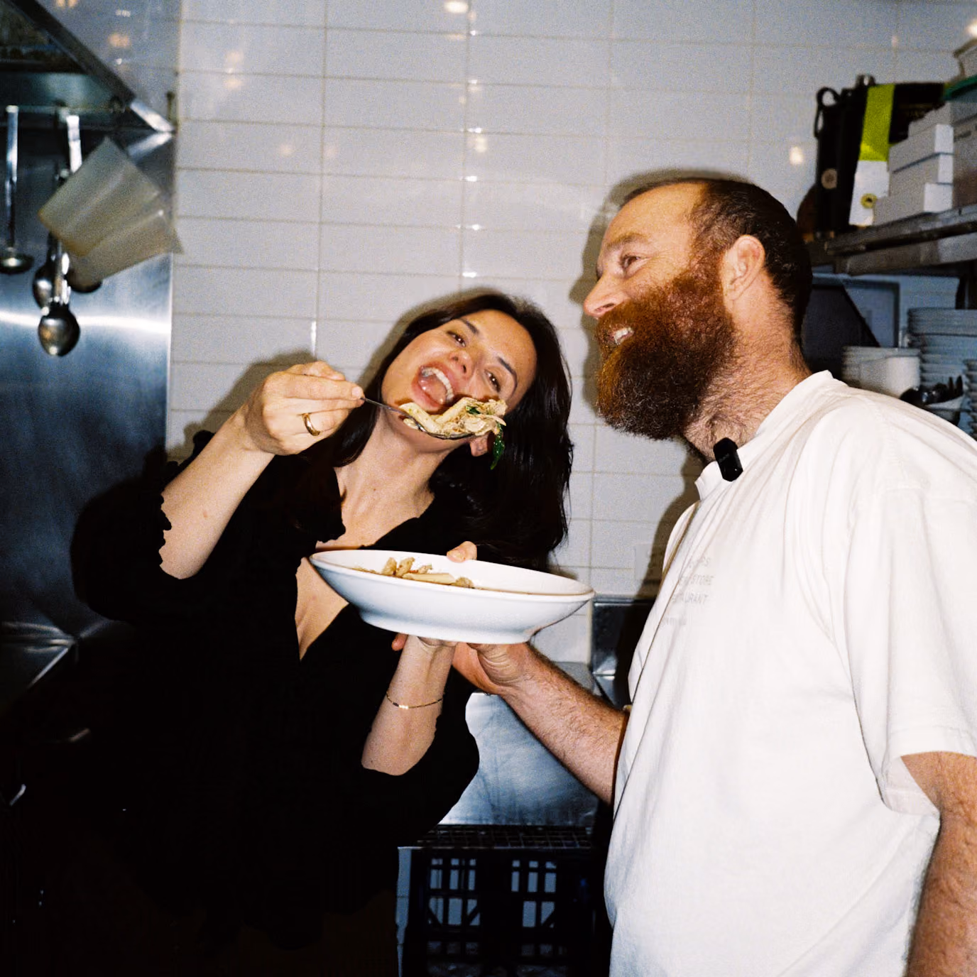 A woman in a black top is about to eat pasta from a fork held near her mouth, while a man with a beard in a white shirt holds a plate of pasta and smiles at her in a kitchen setting.