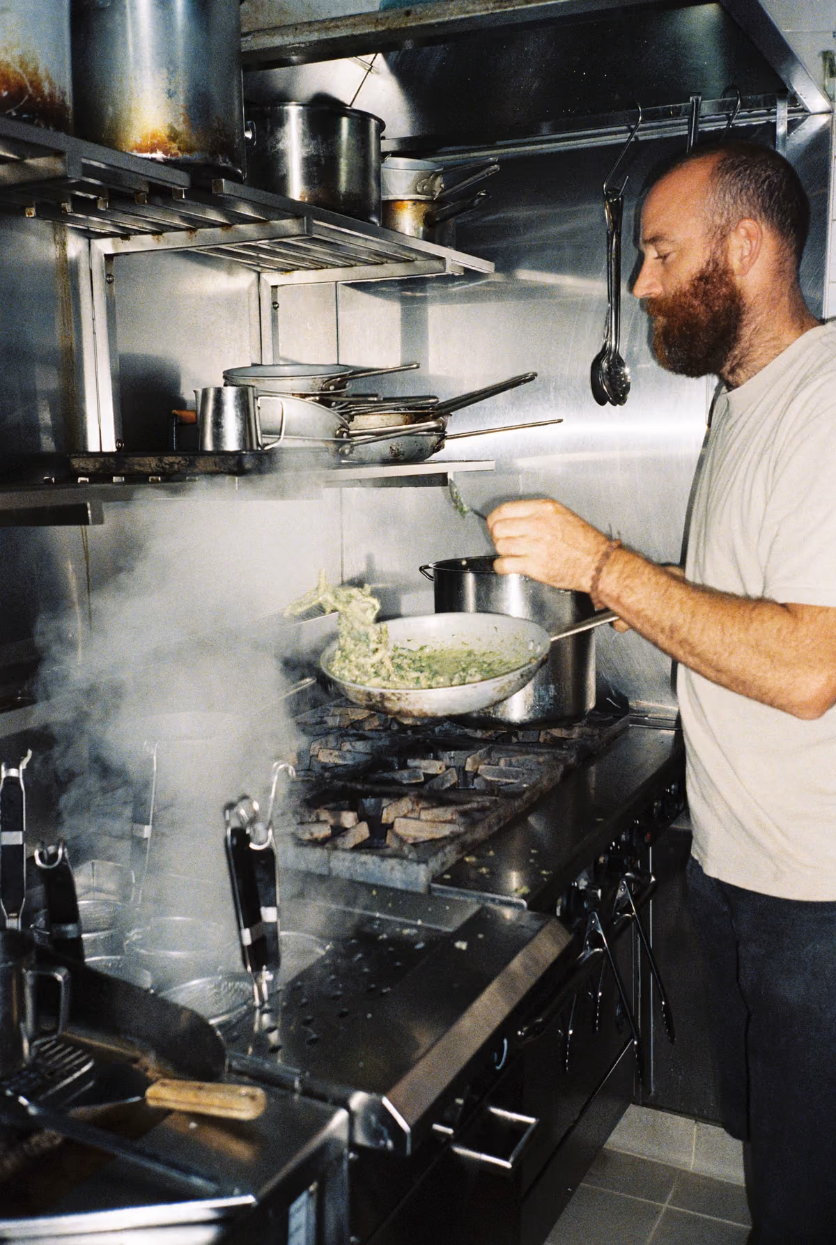 Bearded man cooking pasta in a pan over a commercial stove in a professional kitchen with steam rising.