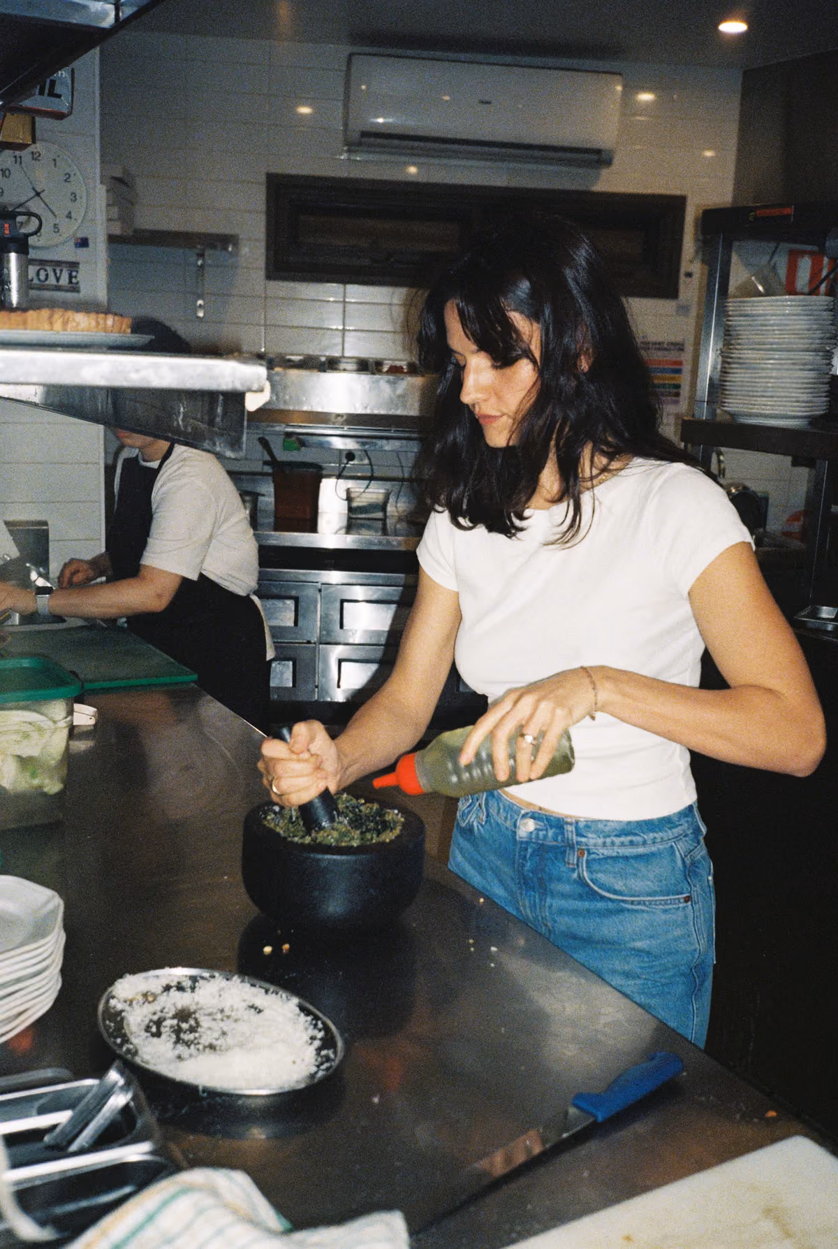A woman in a white t-shirt and blue jeans using a pestle and mortar to crush green herbs in a black bowl on a stainless steel kitchen counter, with kitchen utensils, plates, and another person in the background.