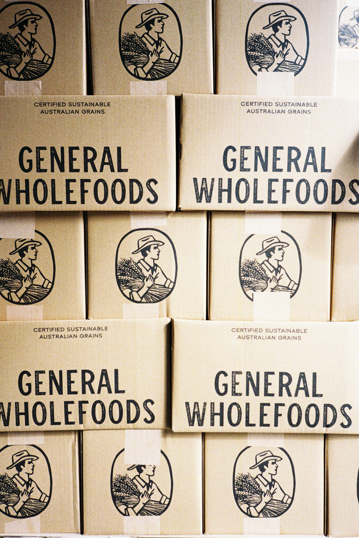 Stack of cardboard boxes labeled 'GENERAL WHOLEFOODS' with a black illustration of a person wearing a hat holding grain bundles and text saying 'CERTIFIED SUSTAINABLE AUSTRALIAN GRAINS'.