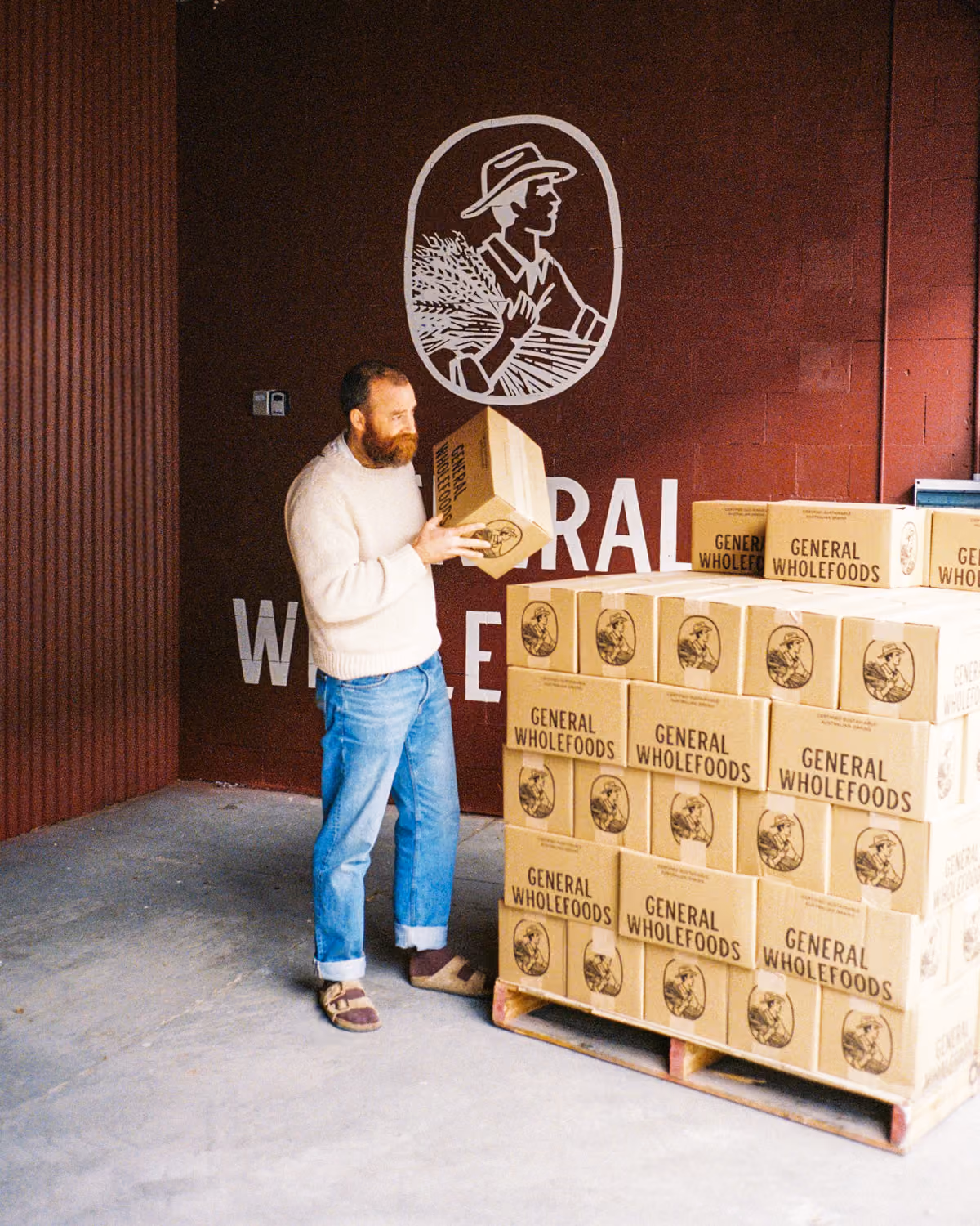 A bearded man in a beige sweater and blue jeans is holding and looking at a cardboard box labeled 'General Wholefoods' in a warehouse next to a pallet stacked with similar boxes, with a dark red wall behind featuring a white logo of a person holding wheat.