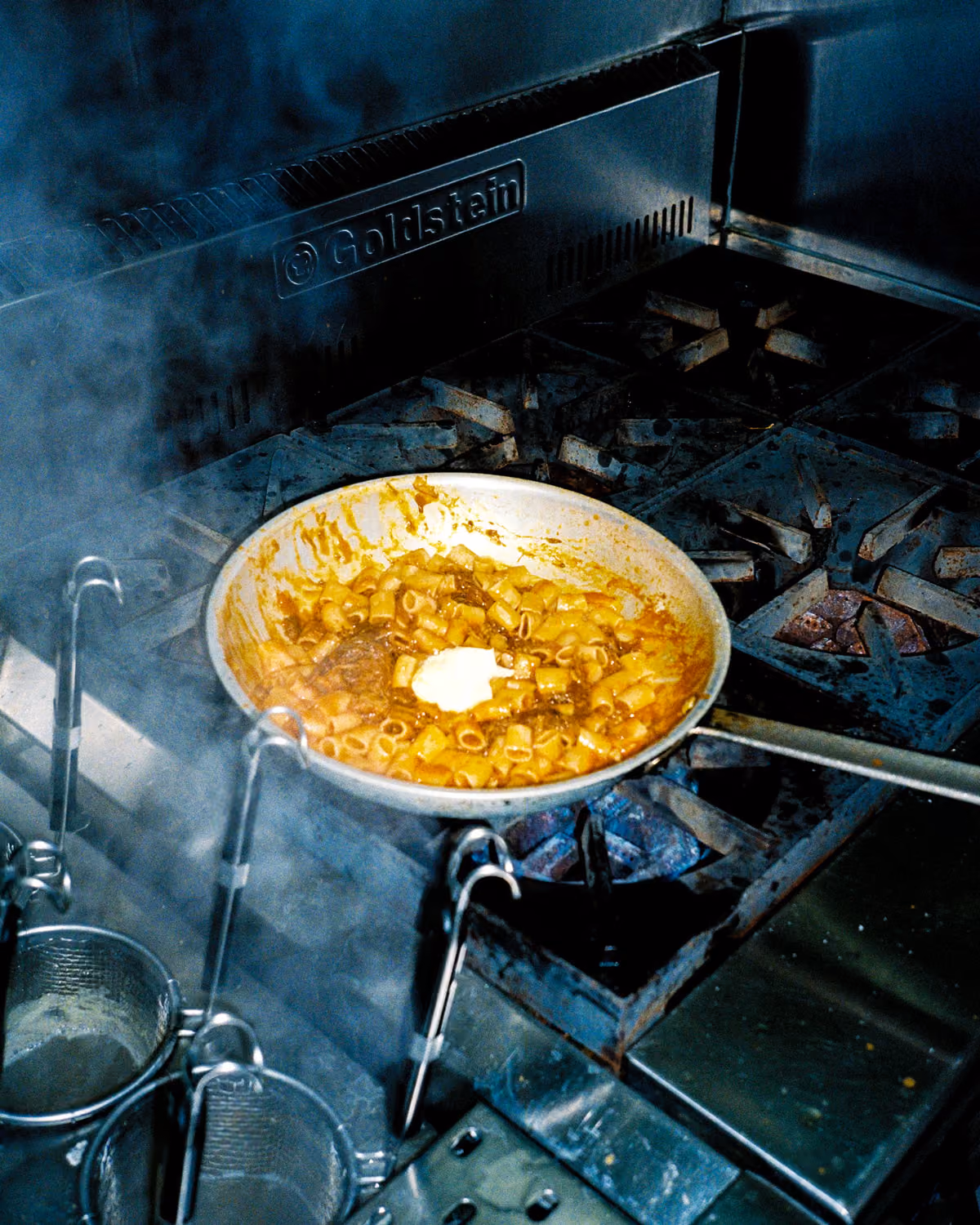 Pan of pasta in a rich sauce with a pat of butter melting on top, cooking on a commercial stove with steam rising around it.