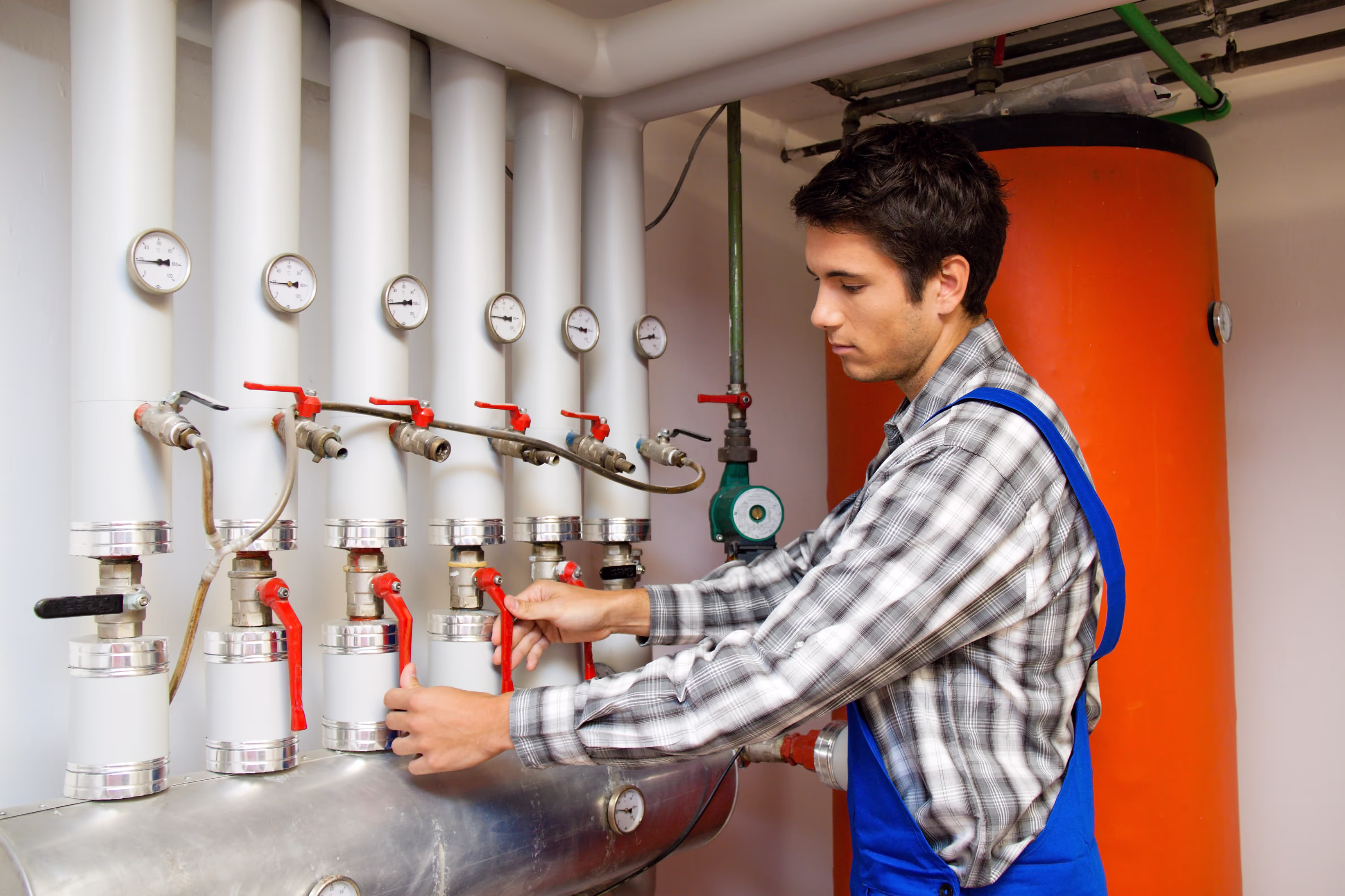 Eine Person arbeitet an einer Heizungsanlage. Sie trägt Arbeitskleidung und betätigt mehrere Ventile und Regler an einem Rohrsystem. Hinter ihr steht ein großer, orangefarbener Warmwasserspeicher. Die Szene wirkt wie eine typische Wartungs‑ oder Installationsarbeit im Heizungs‑ und Sanitärbereich.