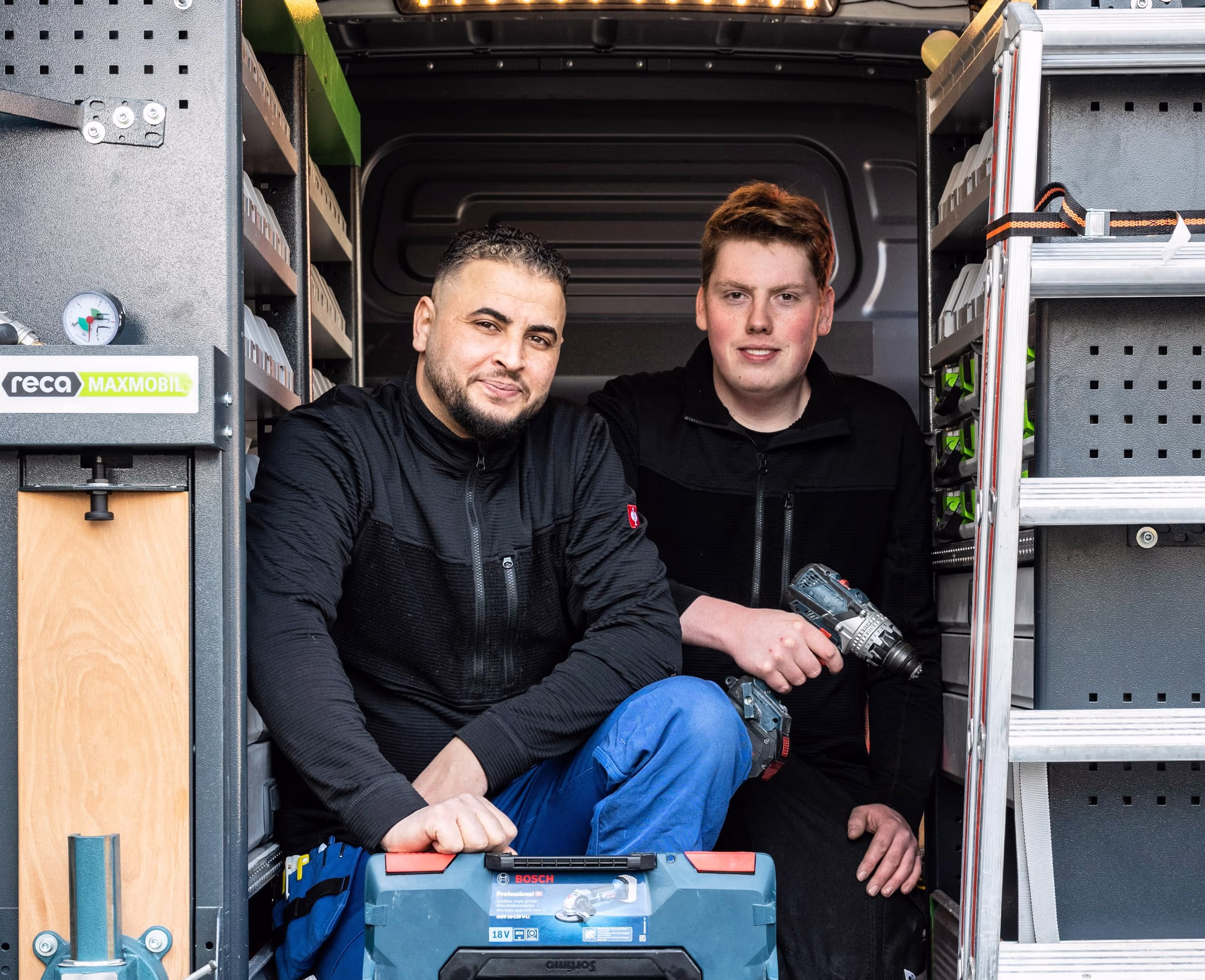 Zwei Personen sitzen im geöffneten Heck eines Einsatzfahrzeugs. Sie tragen Arbeitskleidung, halten Werkzeug (u. a. einen Akkuschrauber) und sind von ordentlich sortierten Regalen mit Ausrüstung umgeben