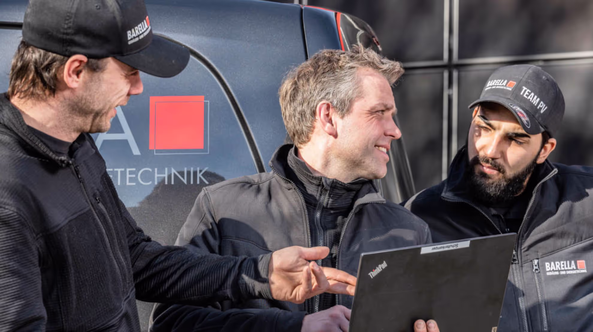 Das Bild zeigt mehrere Personen in Arbeitskleidung, die gemeinsam vor einem Firmenfahrzeug stehen. Eine Person hält ein Laptop, während eine andere darauf zeigt – sie wirken, als würden sie technische Daten besprechen oder eine Aufgabe koordinieren. Alle tragen Outdoor‑ oder Arbeitsjacken und Caps von Barella.