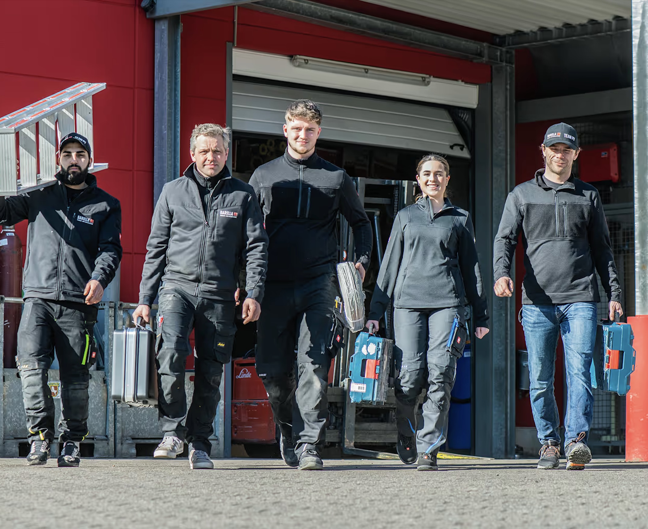Auf dem Foto sieht man fünf Personen, die in Arbeitskleidung selbstbewusst nebeneinander auf die Kamera zugehen. Sie tragen Werkzeugkoffer und wirken wie ein eingespieltes Handwerks‑ oder Serviceteam. Der Hintergrund zeigt ein Gebäude oder eine Werkstatt mit offener Halle.