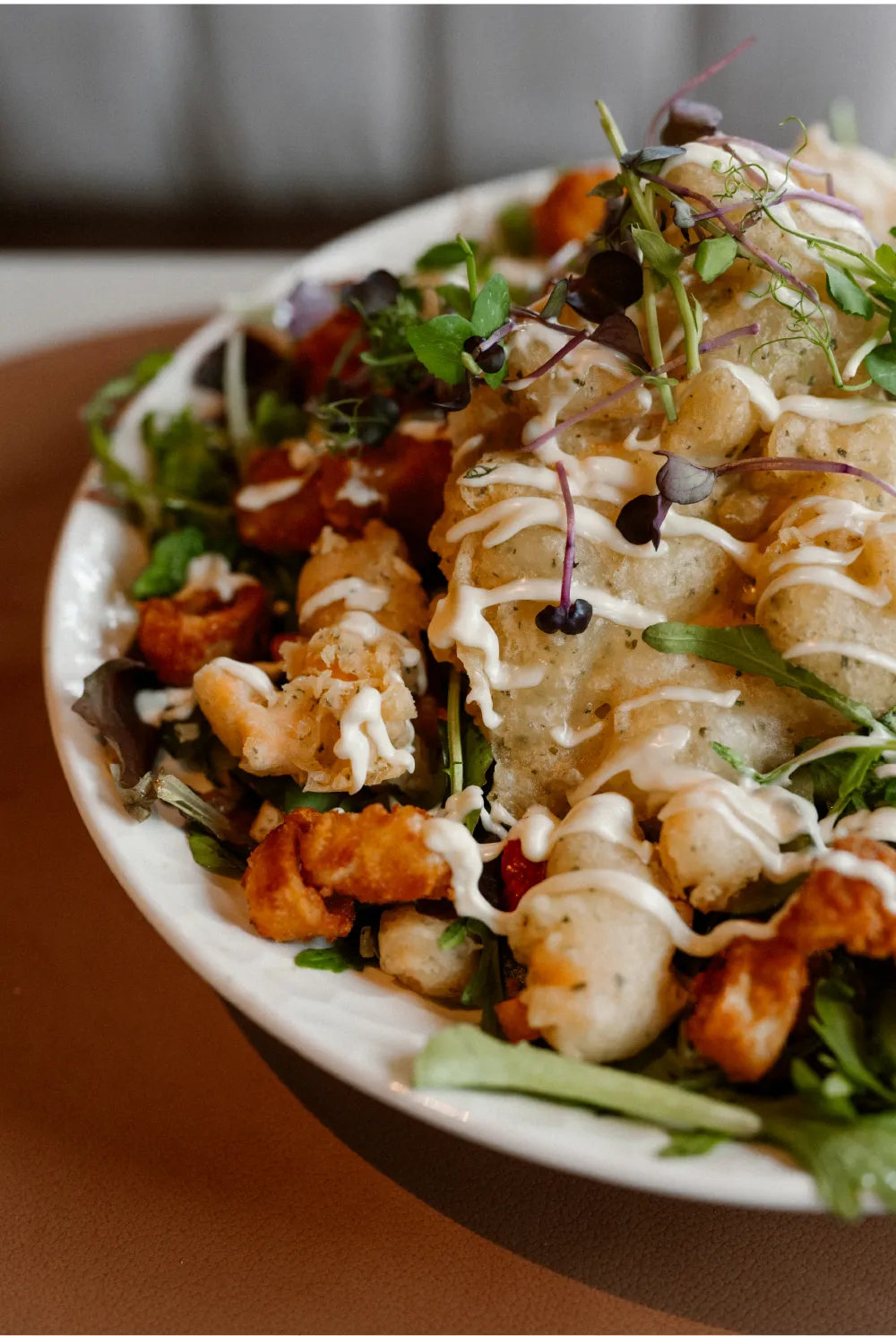 Een bord met een salade met gefrituurde groenten en kip, bedekt met een drizzle van romige dressing en versierd met groene kruiden en microgroenten.