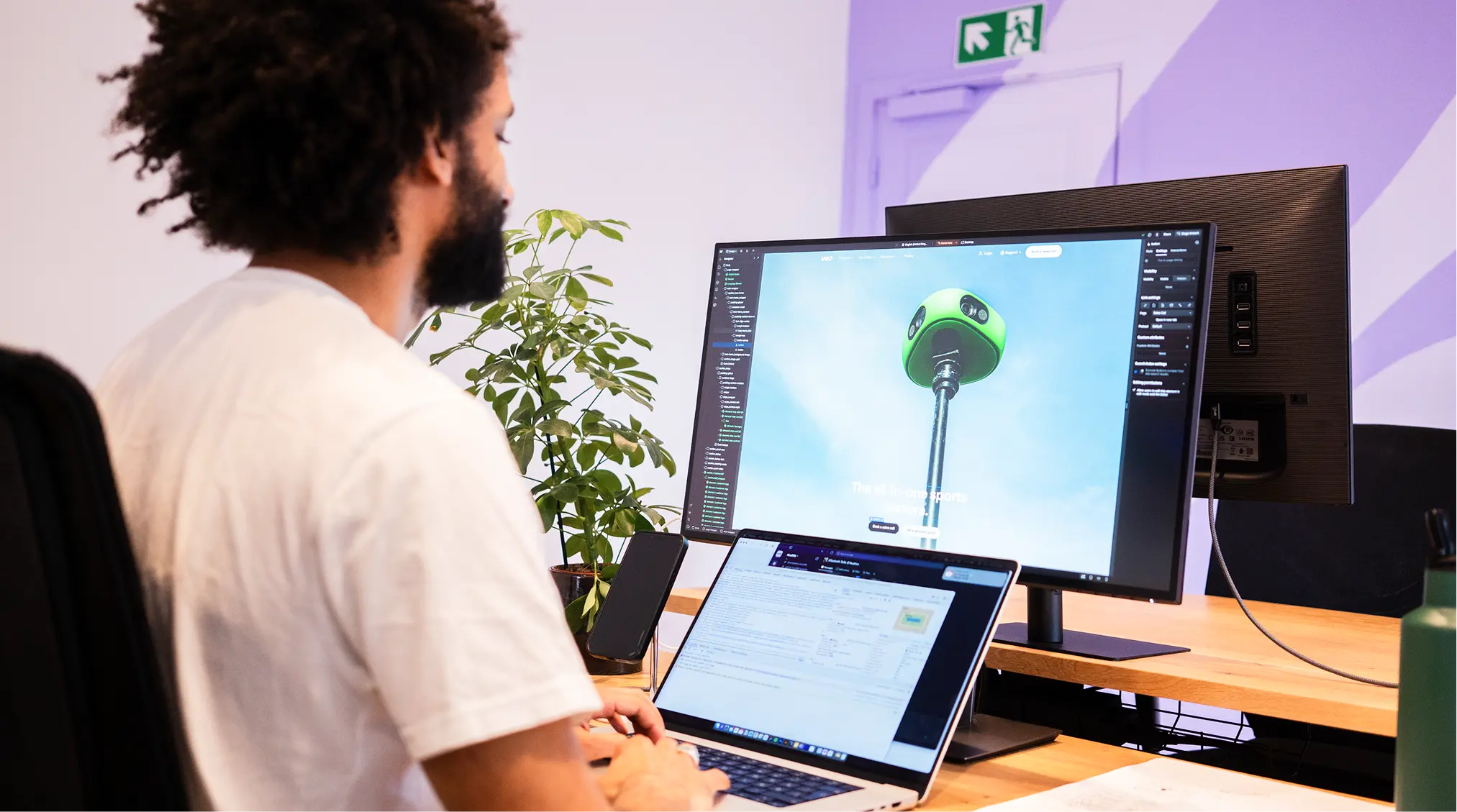 Man with curly hair and beard working on a laptop and a large monitor at a desk with a plant, smartphone, and green water bottle in an office setting.