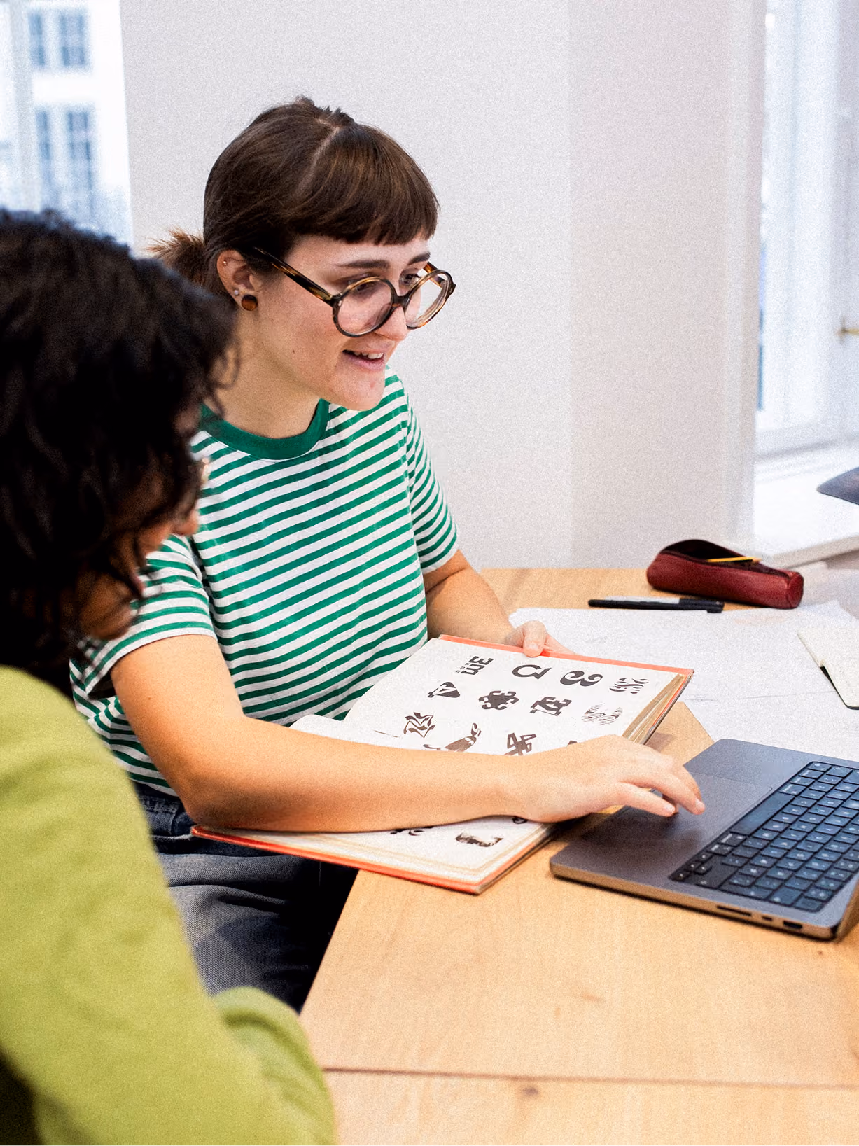 Two people seated at a table collaborating; one person in a green and white striped shirt points at a laptop while holding an open book with graphic designs or logos.