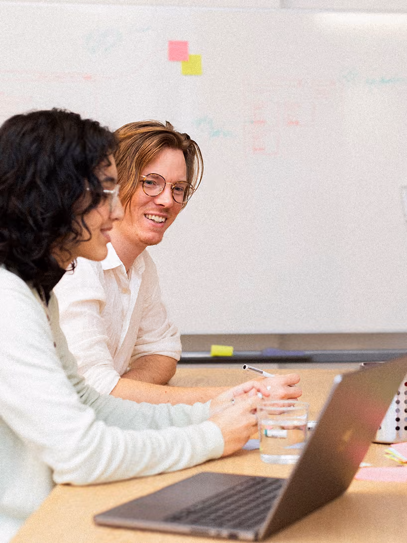 Two people sitting at a desk working together, with one person smiling toward the camera, a laptop in front of them and a whiteboard with sticky notes in the background.