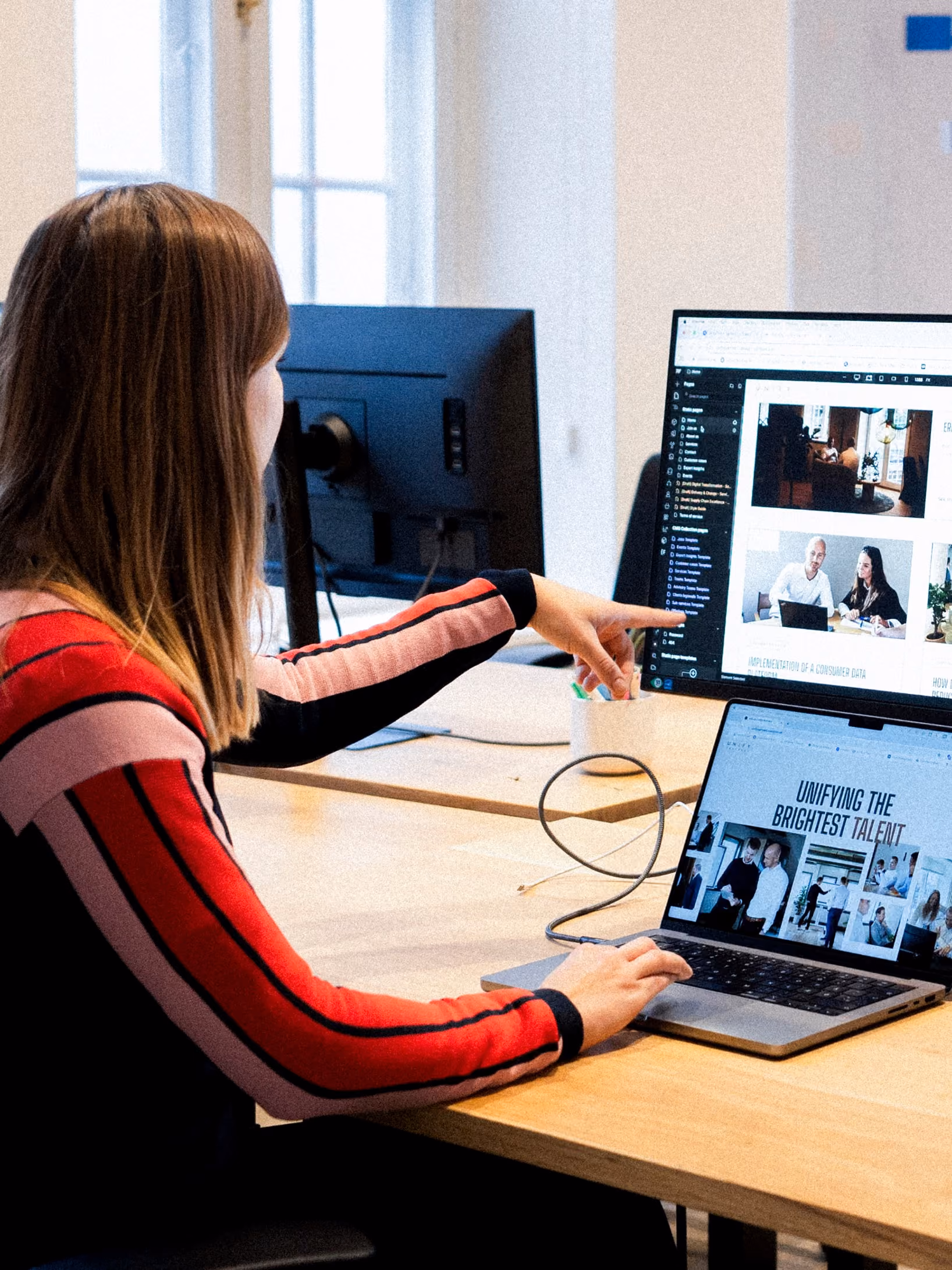 A woman in a red and black striped sweater working at a desk, pointing at a desktop monitor while using a laptop showing a webpage.