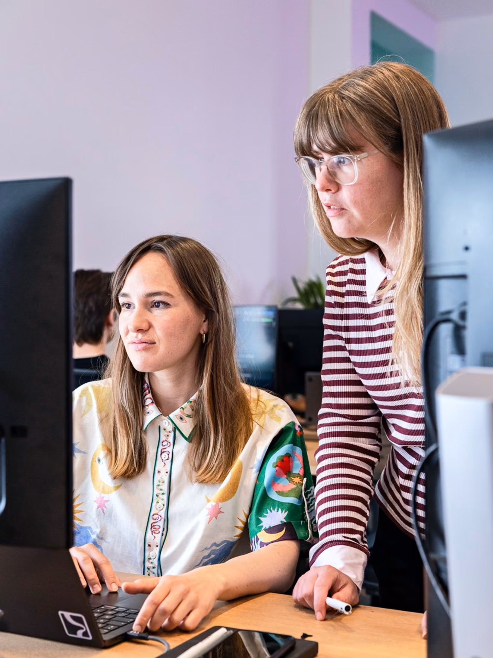 Two women working at desks with computers in an office; the woman sitting wears a colorful patterned shirt and types on a laptop, while the woman standing beside her in a striped shirt looks at the screen.
