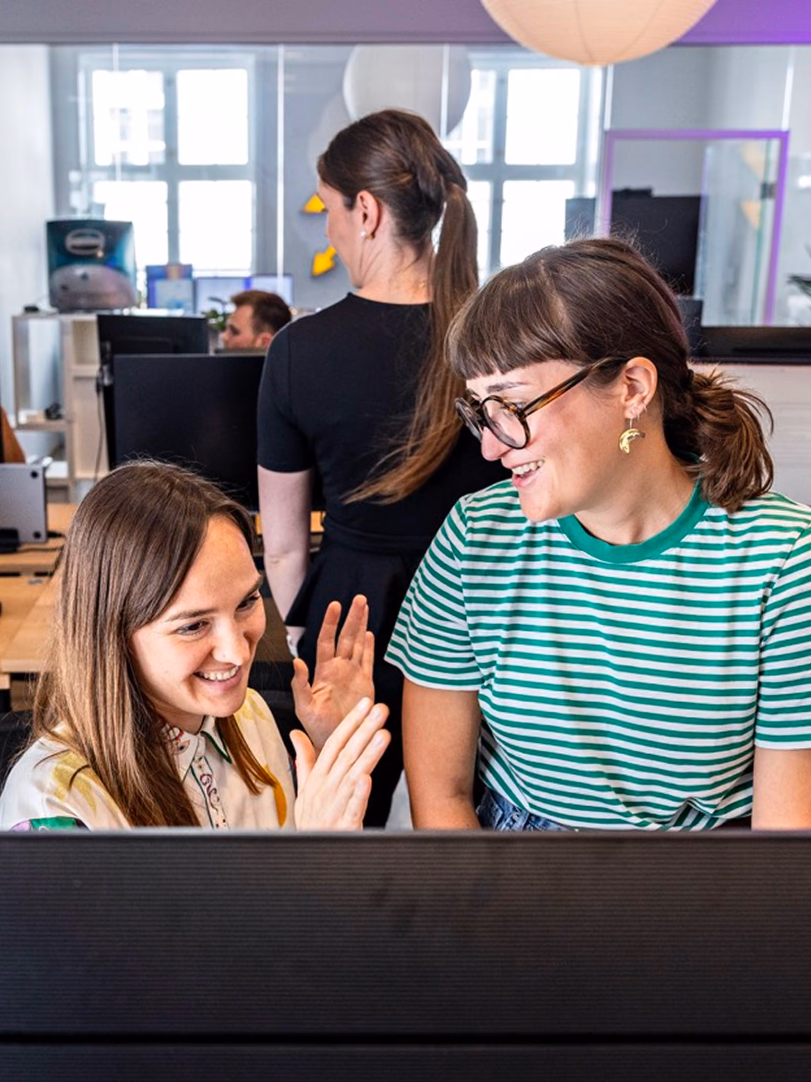 Two women smiling and interacting behind a computer screen in an office environment, with one wearing a green and white striped shirt and glasses, and the other with long brown hair raising her hand.