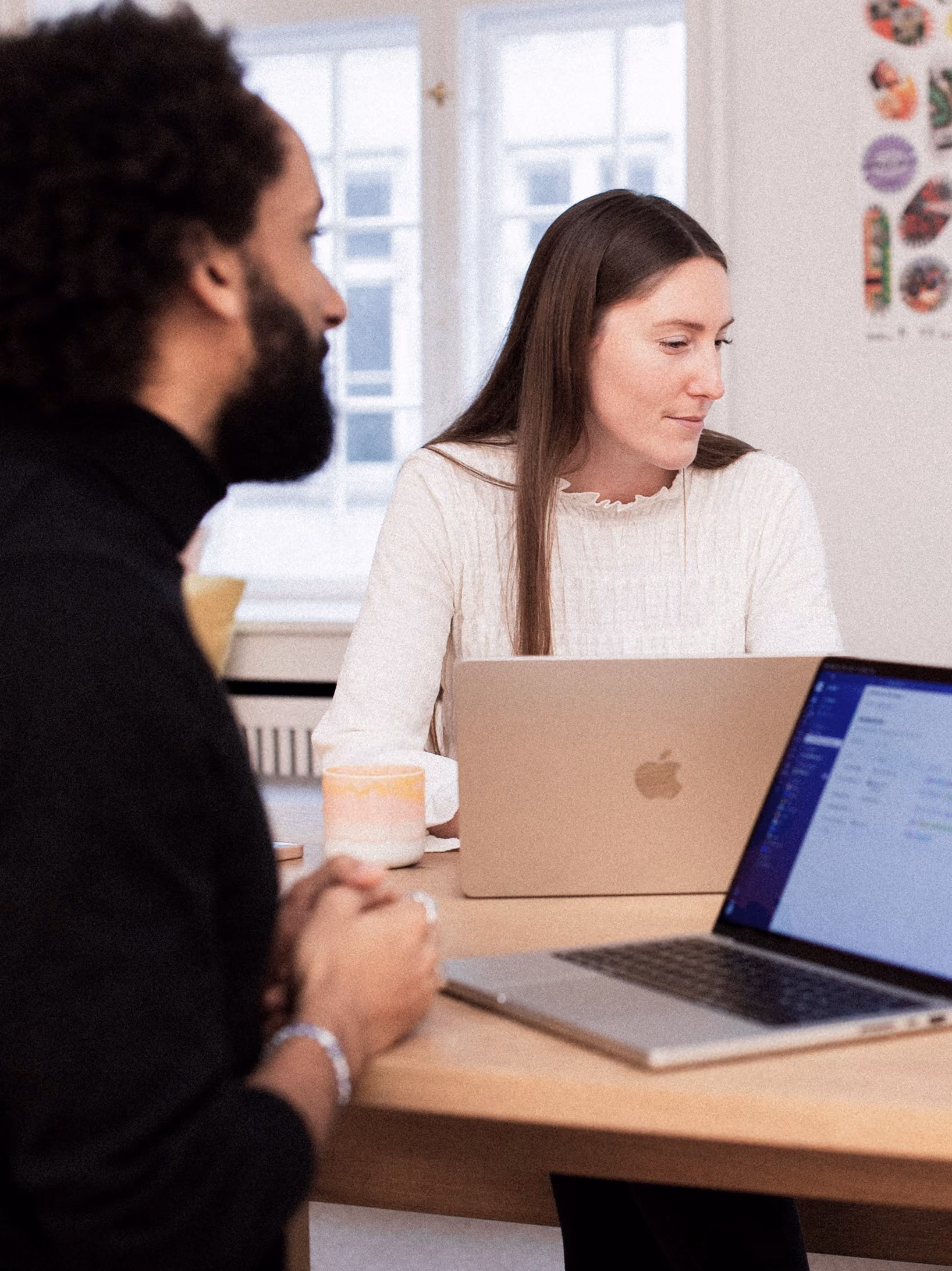 Two people sitting at a wooden table with laptops open, engaged in a discussion in a bright room with windows and wall decorations.