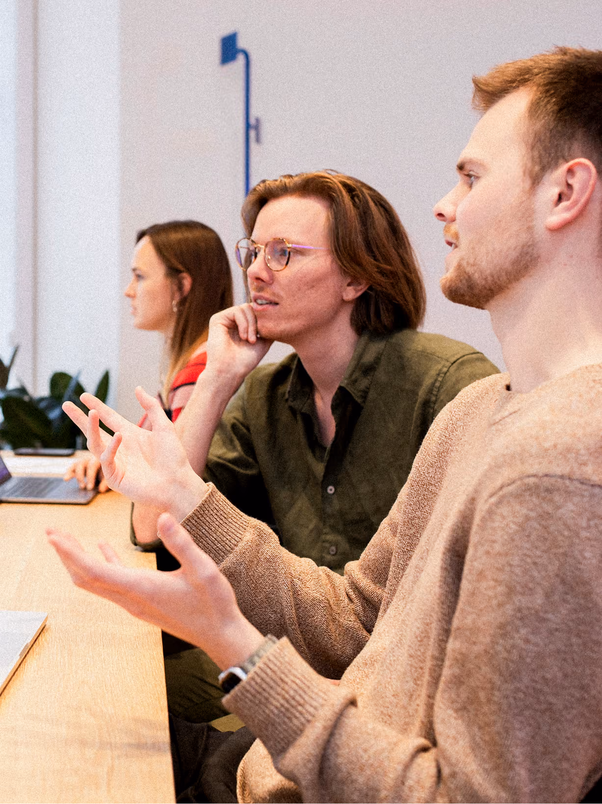 Three people sitting at a table in a discussion, with one person in the foreground gesturing with hands, a second person with glasses listening thoughtfully, and a third person in the background working on a laptop.