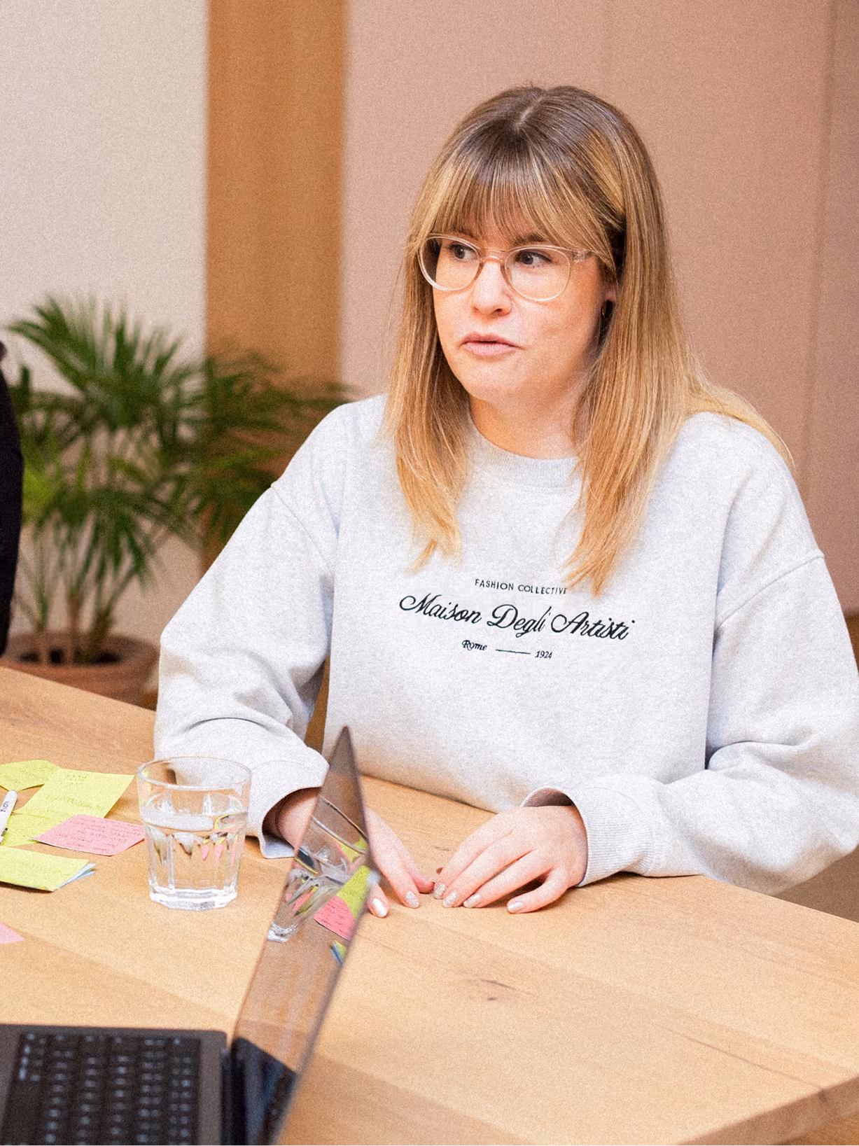 Woman with light brown hair and glasses wearing a gray sweatshirt seated at a wooden table with a glass of water, sticky notes, and a laptop.