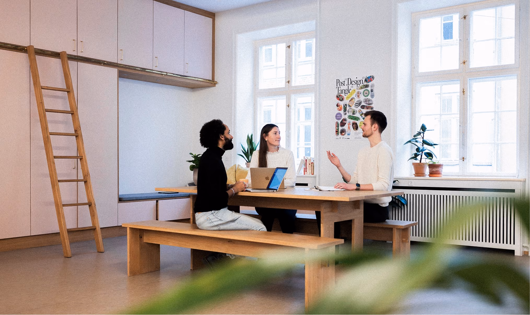 Three people sitting at a wooden table in a bright room with large windows, engaged in conversation with laptops and notebooks in front of them.