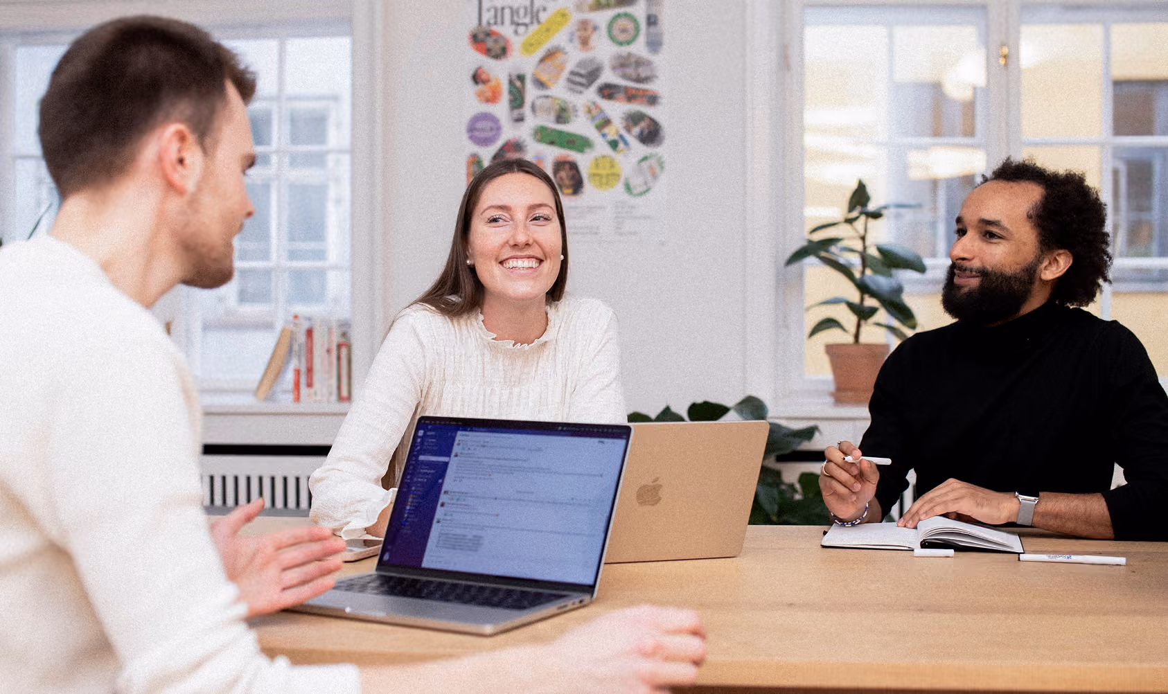 Three diverse young professionals having a meeting around a table with laptops and a notebook, smiling and engaged in discussion in a bright office space with plants and windows.