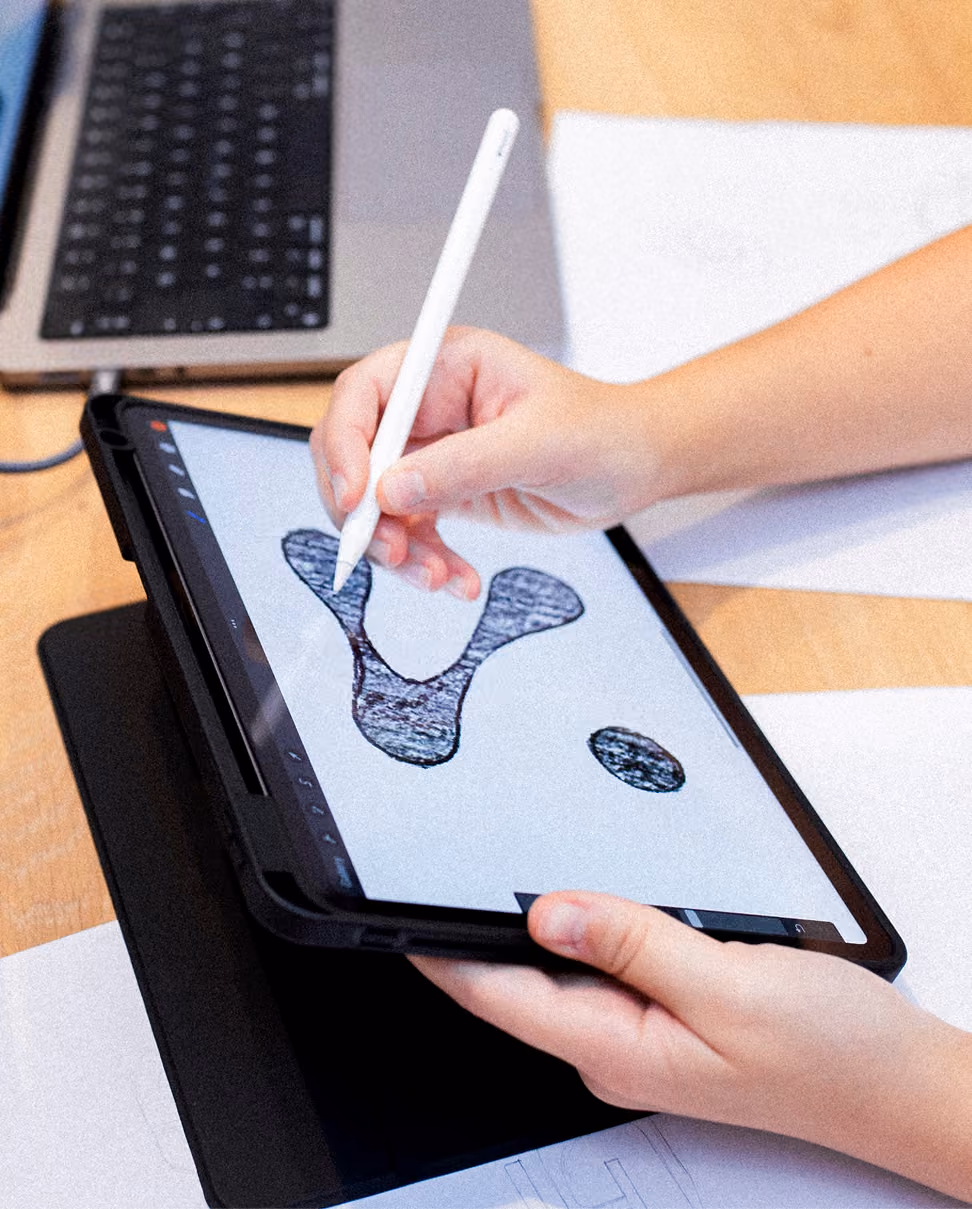 Person using a white stylus to draw a question mark on a tablet screen, with a laptop and papers visible on the wooden desk underneath.