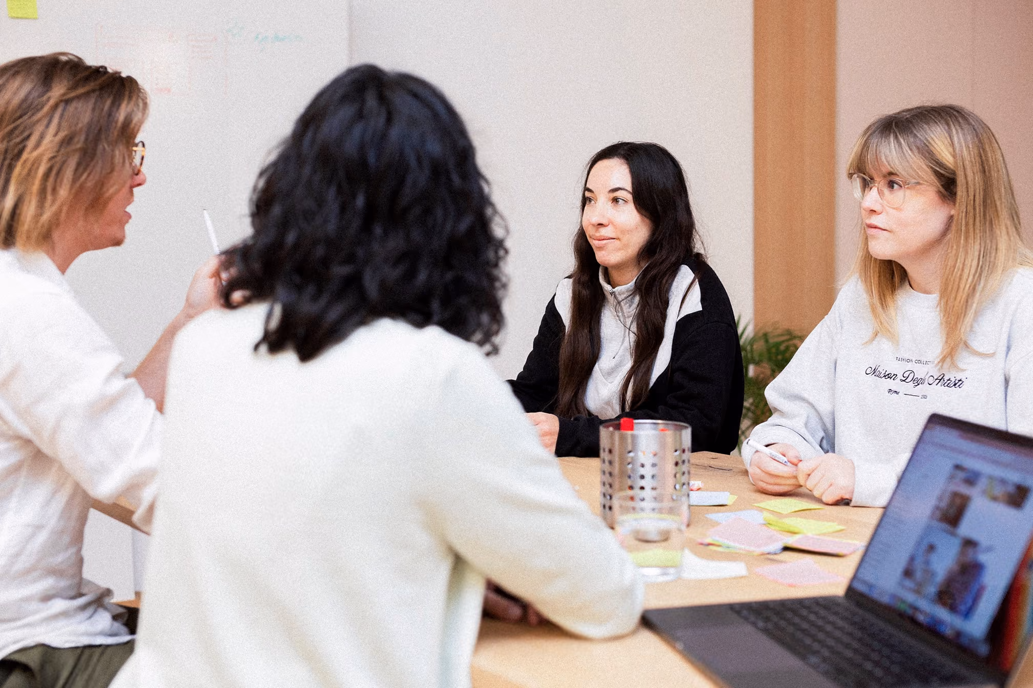 Four women sitting around a table having a discussion in a bright meeting room with sticky notes on the wall and laptop on the table.