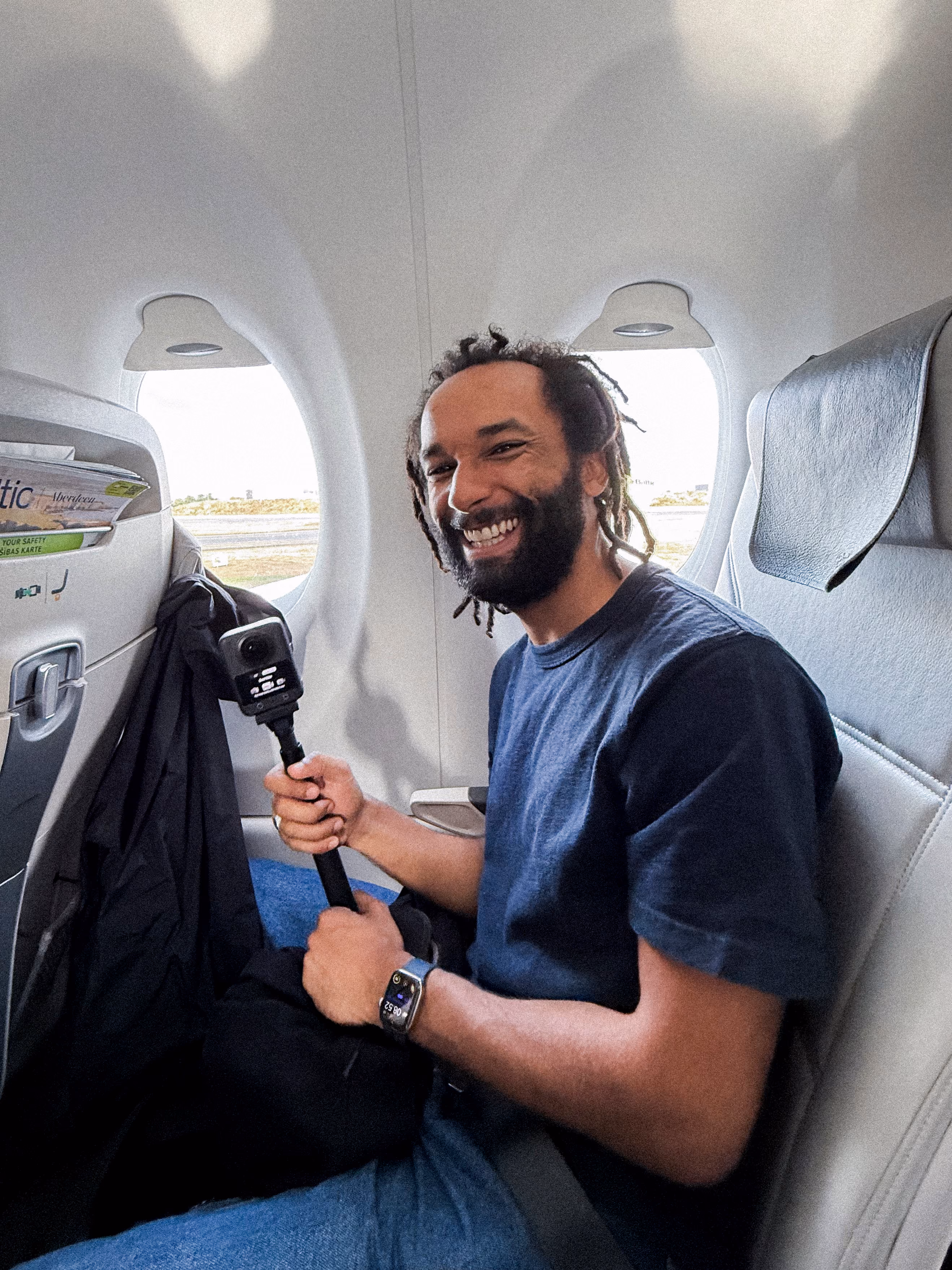 A man with dreadlocks and a beard smiling while sitting in an airplane seat by the window, holding a small camera on a handheld stabilizer, wearing a dark blue shirt and a smartwatch.