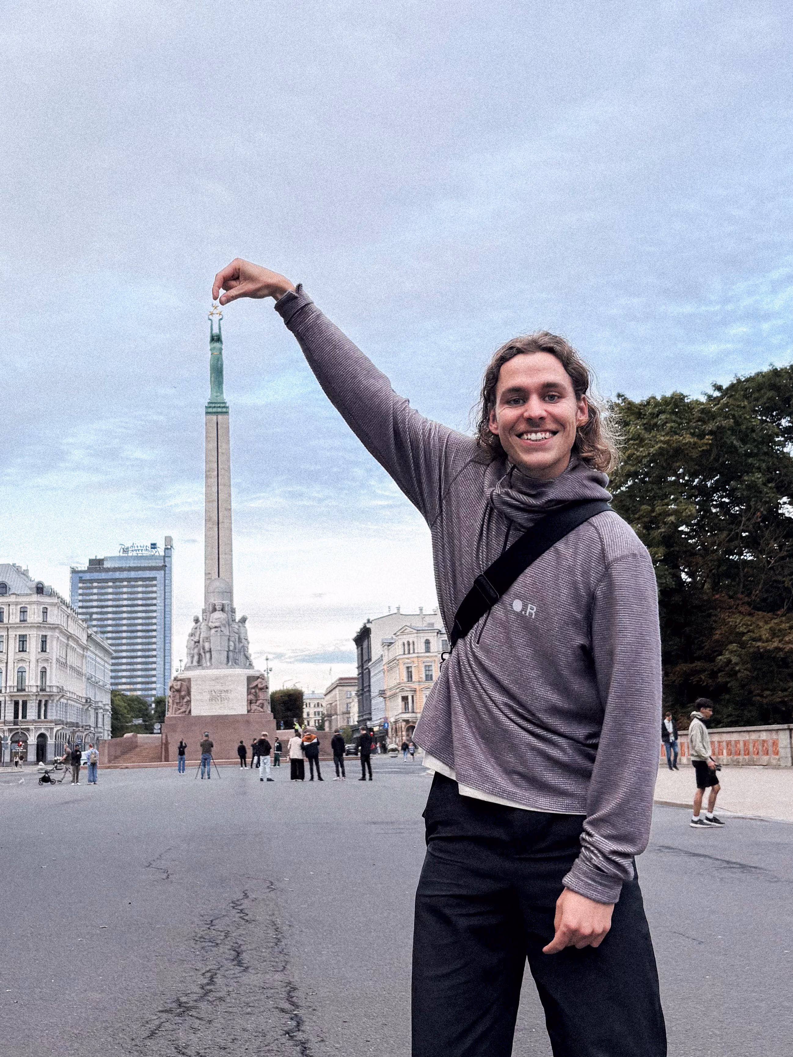 Smiling person wearing a gray hoodie and black pants stands in front of a tall monument, holding their hand up to appear as if pinching the top statue of the monument between their fingers in a public square with other people and buildings in the background.