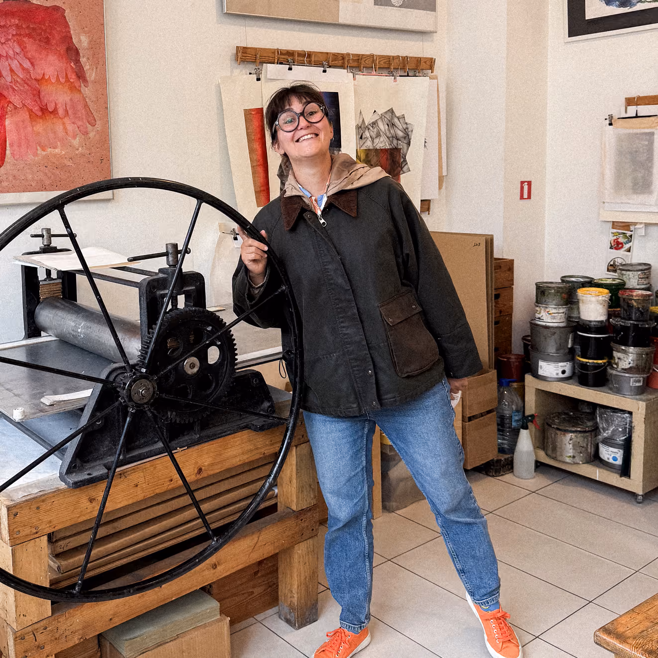 Smiling person with short hair and round glasses wearing a dark jacket and orange sneakers standing beside a large printing press wheel in an art studio with paint cans and drawings on the walls.