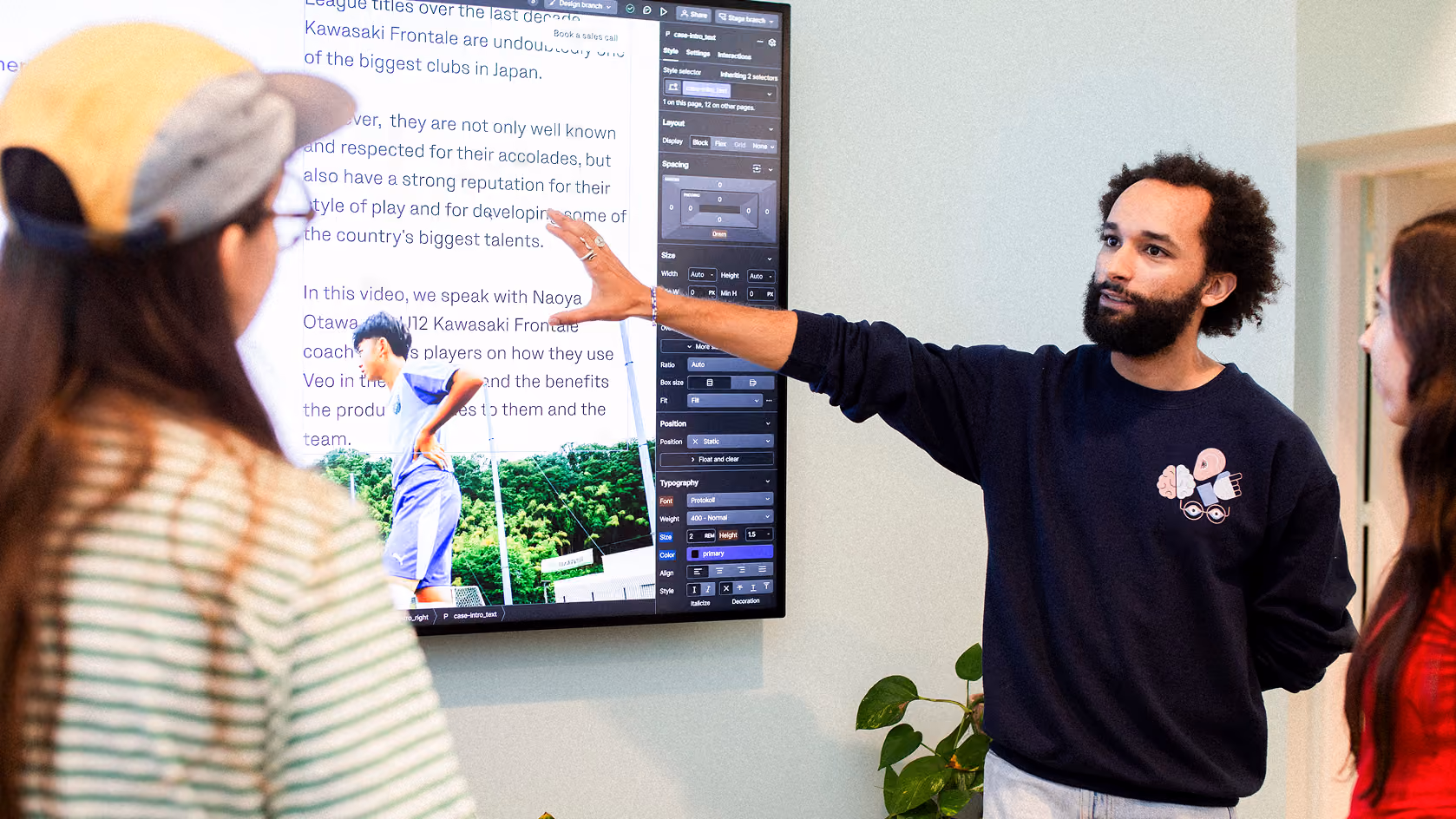 Man with curly hair and beard pointing at a large screen displaying text and an image of a soccer player, while two women look on attentively in a casual meeting setting.