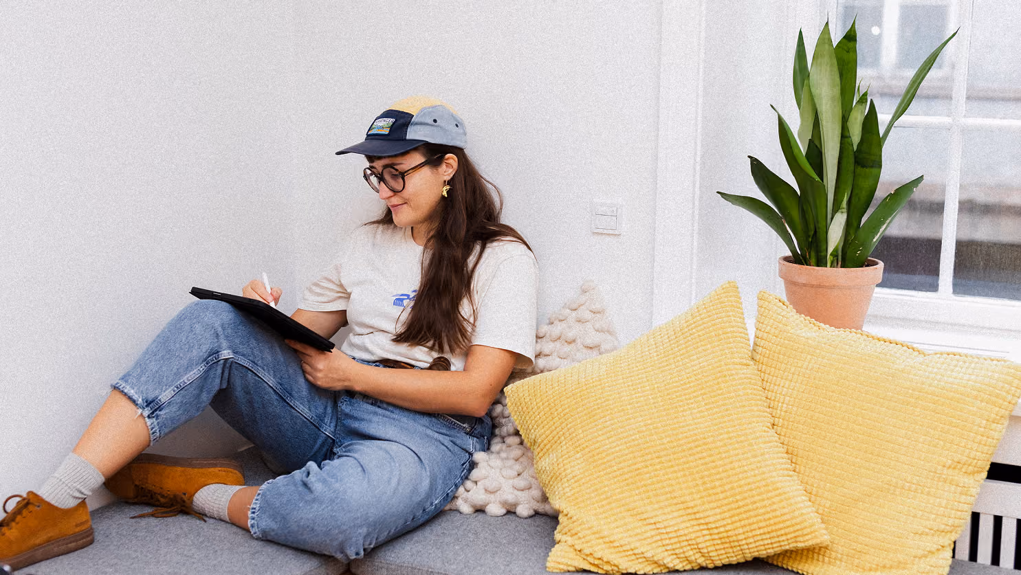 A young woman wearing a colorful cap, glasses, beige t-shirt, and blue jeans sits on a cushioned bench with yellow pillows, writing on a black tablet with a stylus, near a houseplant and window in a bright room.