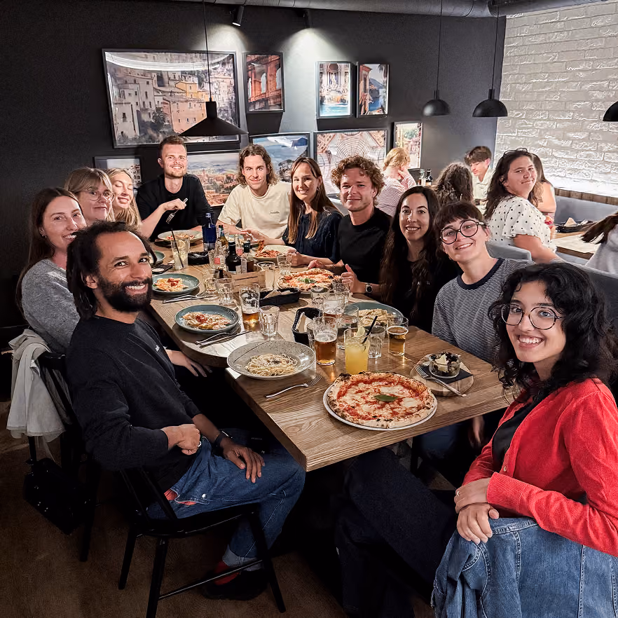 Group of twelve people sitting around a wooden table in a restaurant, smiling and enjoying various dishes including pizza and pasta, with drinks and framed pictures on the dark walls behind them.