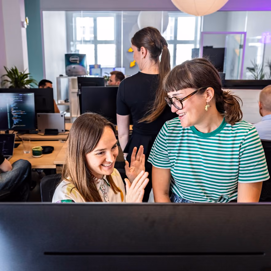 Two women smiling and engaging in conversation at a desk with a large computer monitor in front of them in a busy modern office.