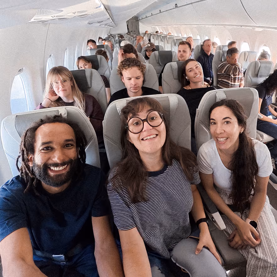 Diverse group of passengers seated inside an airplane cabin with three people in the front row smiling at the camera.