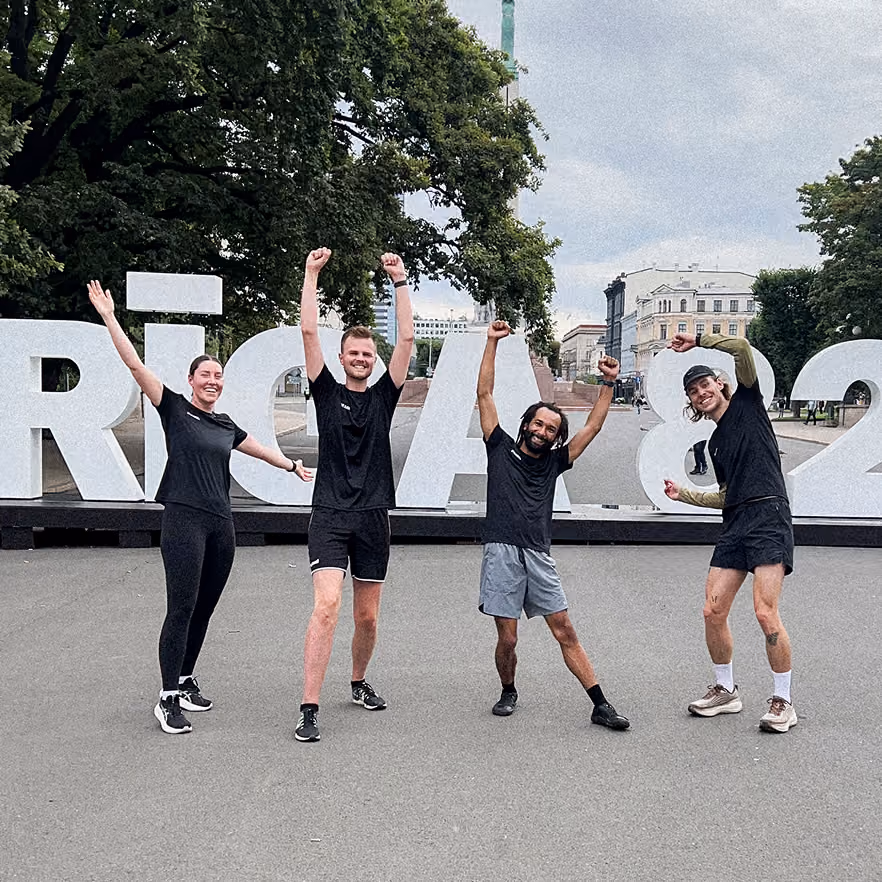 Four people standing outdoors in front of large white letters on a platform, posing with raised arms and smiling, wearing casual athletic clothing and sneakers, with trees and buildings in the background under a cloudy sky.