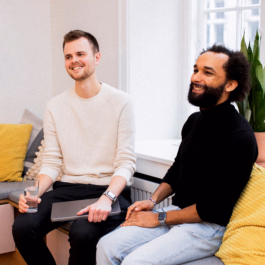 Two men sitting on a couch in a bright room; the man on the left wears a white sweater and black pants, holding a glass of water and laptop, the man on the right wears a black turtleneck and light jeans, both smiling.
