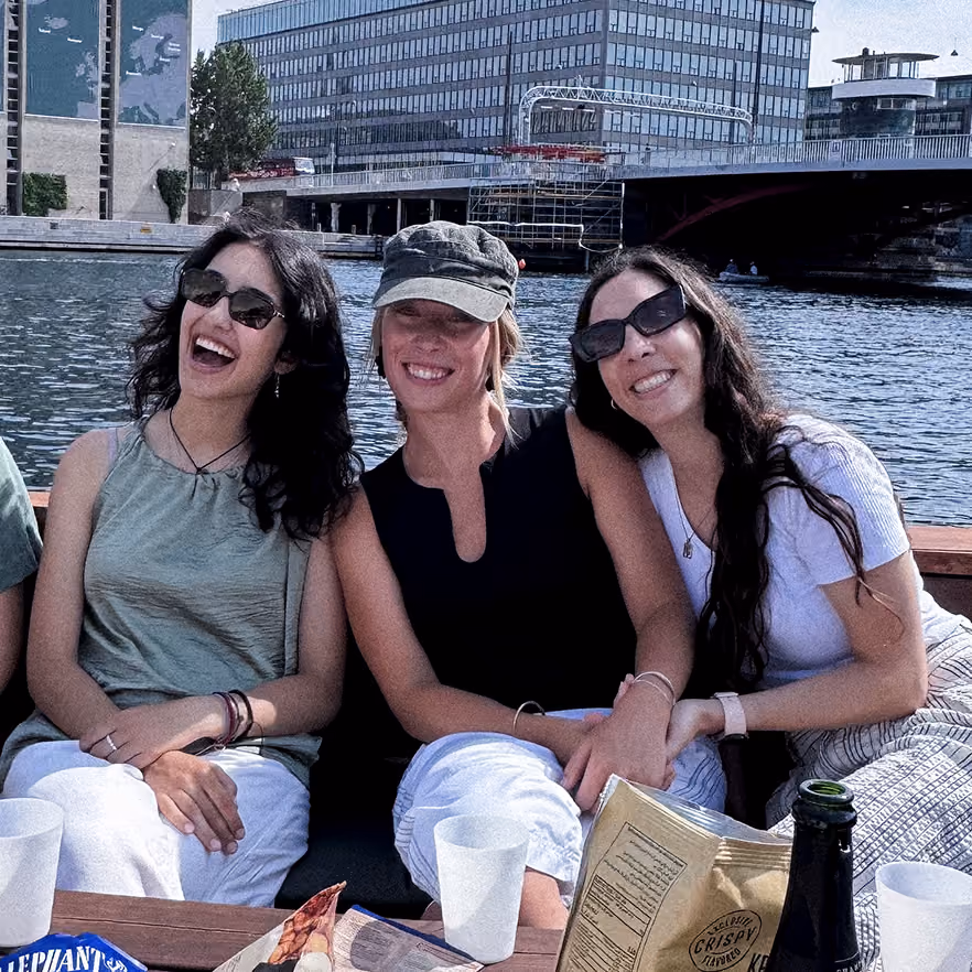 Three smiling women wearing sunglasses sit closely on outdoor bench by river with buildings and bridge in background, sharing snacks on table in front.