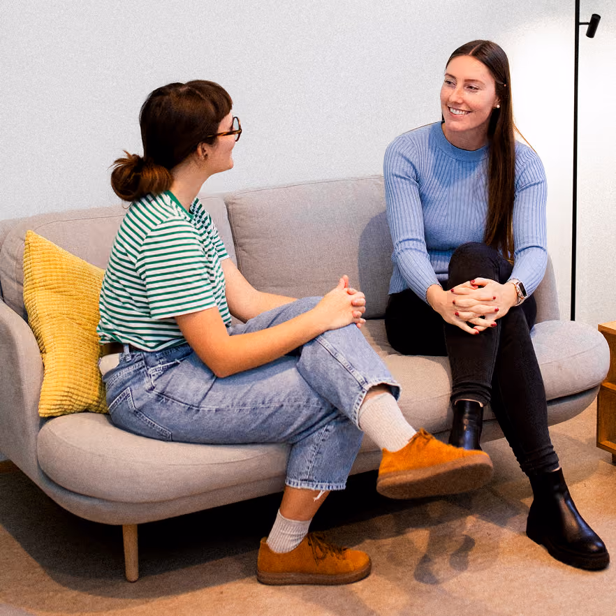 Two women sitting and chatting on a light gray couch indoors, one wearing a green and white striped shirt and jeans with brown shoes, the other wearing a blue sweater, black pants, and black boots, with a yellow pillow on the couch beside them.