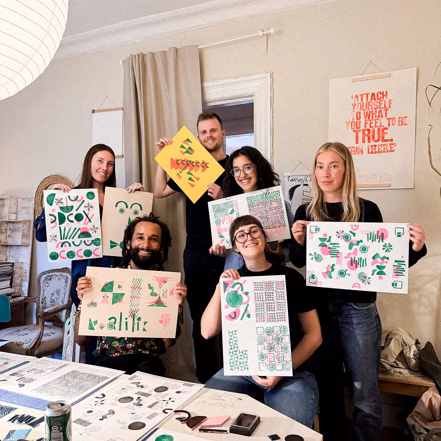 Group of six smiling people in a room holding up colorful abstract artwork consisting of geometric shapes and patterns in green, pink, and yellow, with artwork spread on the table in front of them.