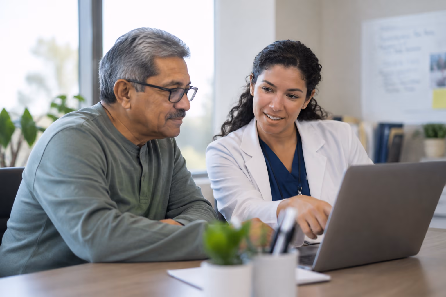 A female doctor in a white coat is sitting at a desk, pointing to a laptop screen while explaining something to an older man wearing glasses and a green sweater.