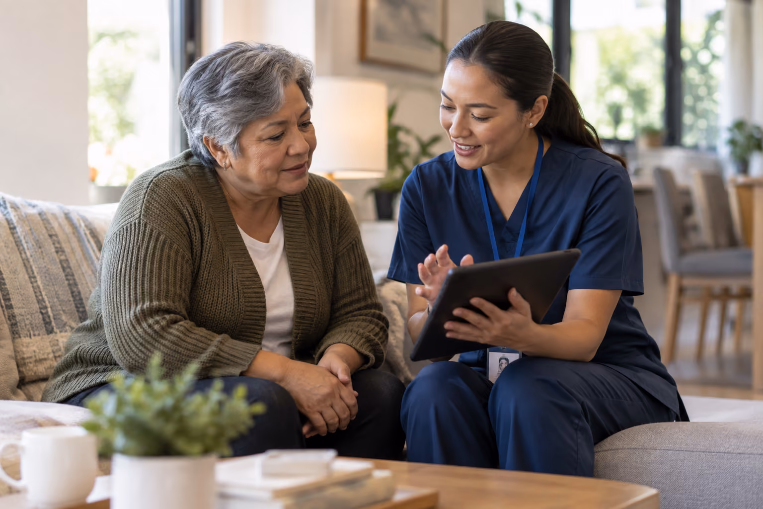 A young female healthcare worker in blue scrubs sitting on a couch and showing information on a tablet to an older woman in a green cardigan inside a bright living room.