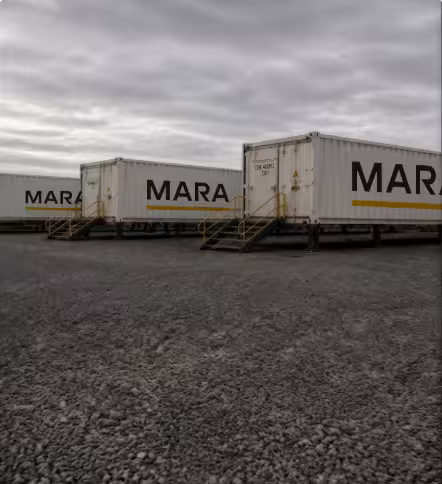Row of white MARA branded modular mining containers elevated on stilts with yellow-railed access stairs across a gravel pad.