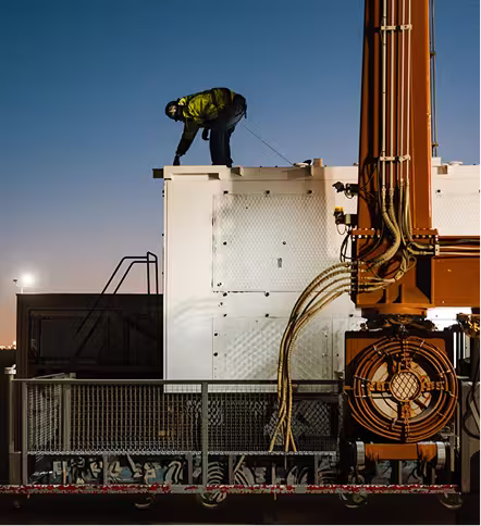 Technician in high-visibility safety gear working on top of a white modular mining container beside heavy industrial equipment.