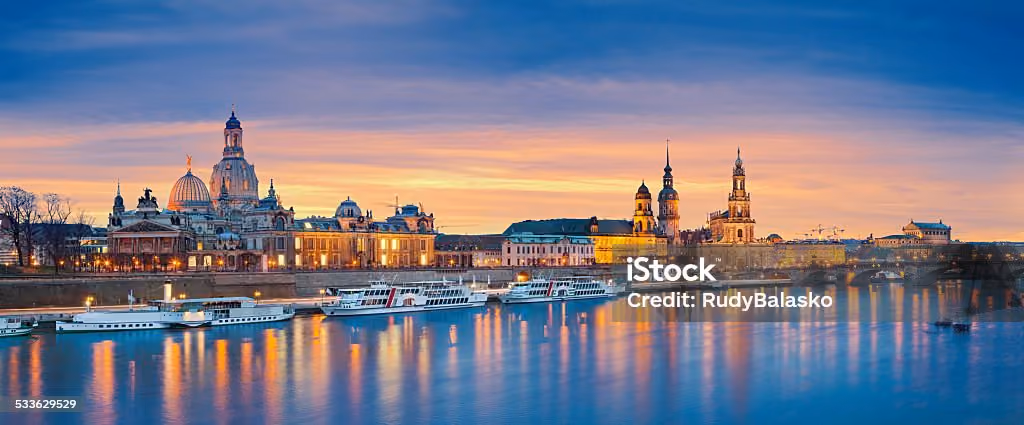 Panoramablick auf die historische Dresdner Altstadt bei Sonnenuntergang mit beleuchteten Gebäuden und Kirchtürmen, die sich in der Elbe spiegeln.