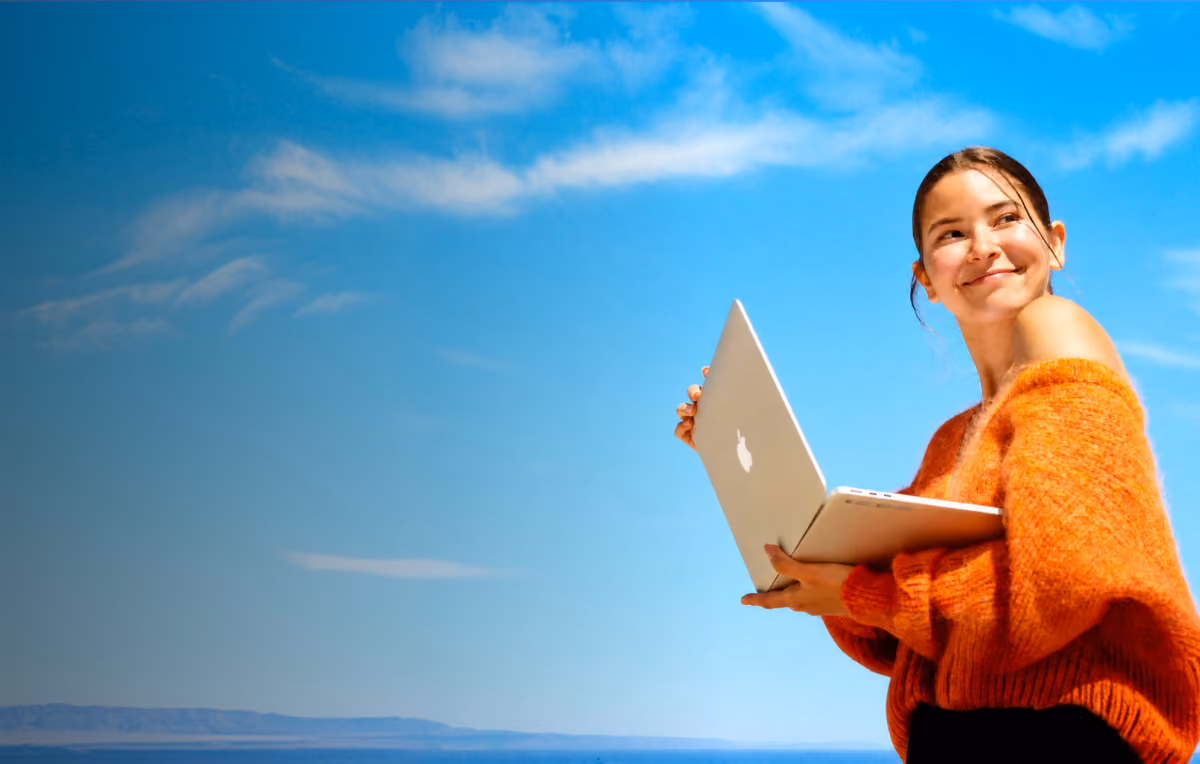 Young woman wearing an orange sweater holding a white laptop outdoors with a blue sky and distant hills in the background.