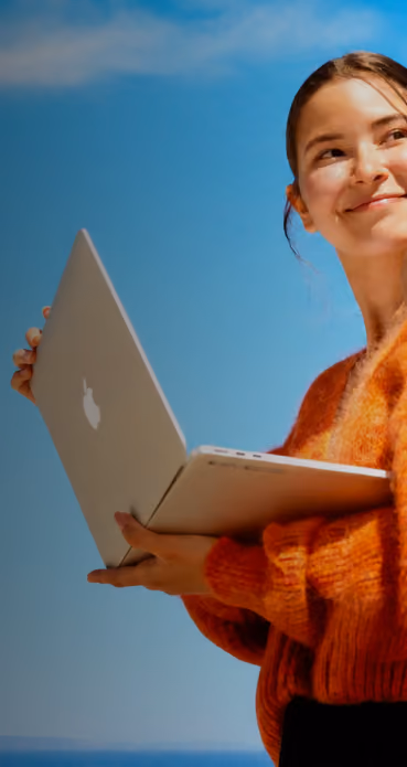 Young woman outdoors holding an open silver MacBook laptop, smiling and looking to her right against a clear blue sky.