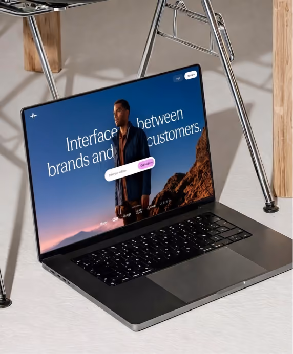 Open laptop on a white surface under a metal chair frame, displaying a website homepage with a man standing outdoors and text that reads 'Interface between brands and customers.'