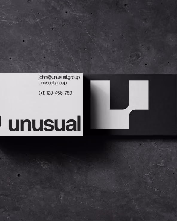 Two business cards on dark textured surface; left card white with contact email john@unusual.group, website unusual.group, phone (+1) 123-456-789, and bold black text 'unusual' partially visible, right card black with abstract light gray geometric logo.